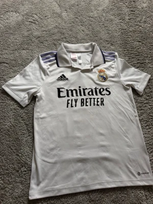 Real Madrid matchtröja Adidas vit - Snygg vit matchtröja från Real Madrid med broderat klubbmärke och Adidas-logga. Tröjan har korta ärmar, krage med svart och lila detaljer samt tre klassiska ränder på axlarna. Tillverkad i lätt och ventilerande material, perfekt för fotboll eller träning.