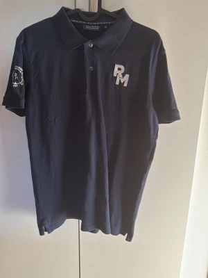 Marinblå piké från Race Marine - Snygg marinblå pikétröja från Race Marine med broderad RM-logga på bröstet och patch på ärmen. Klassisk krage och knappar framtill. Perfekt för en sportig och avslappnad stil.