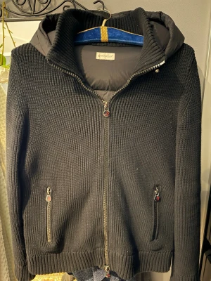Moncler cardigan (äkta) - Svart pufferjacka från Moncler med stickade ärmar och huva. Jackan har två fickor med dragkedja och ribbad kant vid ärmslut och nederkant. Framsidan har en tydlig Moncler-logga och jackan är quiltad på ryggen och huvan. Perfekt för kyliga dagar. (Inte säker men troligen ÄKTA)