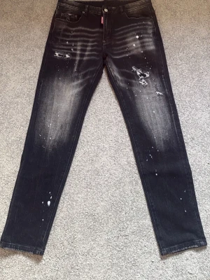 Dsquared jeans   - Säljer ett par svarta jeans med slitningar och vita färgstänk för en riktigt edgy look. Jeansen har raka ben, klassisk femficksmodell och coolt tvättad finish. Perfekta för dig som gillar streetwear och vill sticka ut lite extra.