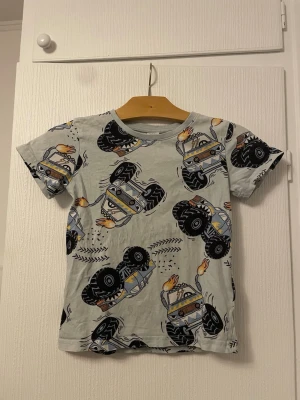 T-shirt med monstertruckar från Lindex - Cool ljusgrå t-shirt från Lindex med färgglatt monstertruck-motiv över hela plagget. T-shirten har rund halsringning och korta ärmar. Perfekt för dig som gillar bilar och vill sticka ut med ett lekfullt tryck.