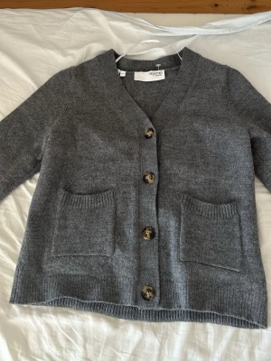 Grå kofta med knappar från Selected - Mysig grå kofta från Selected med stora knappar framtill och två praktiska fickor. Koftan har v-ringning, ribbade muddar och är tillverkad i ett mjukt stickat material. Koftan är i storlek M men har krympt något i tvätten och passar mer som S, XS.