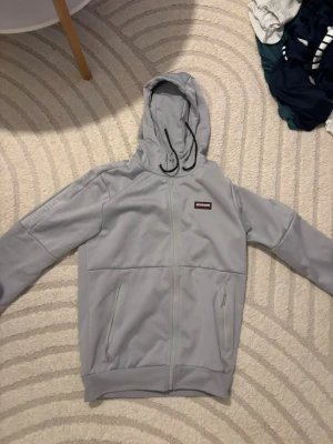 Ljusgrå hoodie från McKenzie - Snygg ljusgrå hoodie från McKenzie med dragkedja framtill och två fickor med dragkedja. Huva med justerbar dragsko och diskret logga på bröstet. Perfekt för chill dagar eller när du vill ha en enkel och clean look.