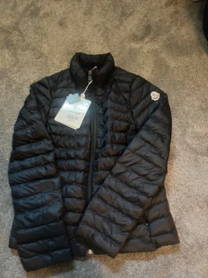 Svart dunjacka från Moncler - Snygg svart dunjacka från Moncler med quiltad design och hög krage. Jackan har dragkedja framtill och en Moncler-logga på ärmen. Perfekt för kalla vinterdagar och riktigt lätt att bära. Klassisk och stilren modell som passar till det mesta.