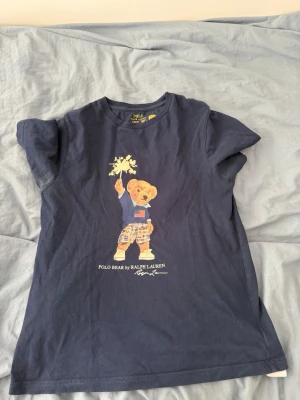 Marinblå Polo Bear t-shirt Ralph Lauren - Snygg marinblå t-shirt från Ralph Lauren med den ikoniska Polo Bear-trycket framtill. T-shirten har rund hals, korta ärmar och är gjord i mjuk bomull. Perfekt för en avslappnad och cool stil.