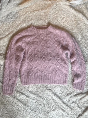 Ljuslila kabelstickad tröja - Supersöt ljuslila stickad tröja med kabelstickat mönster på framsidan och ärmarna. Tröjan har rund hals, ribbade muddar och är gjord i ett fluffigt, mjukt material som känns riktigt skönt. Perfekt att matcha med jeans eller kjol för en mysig look.