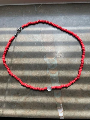 Rött halsband med pärlor - Säljer ett handgjort halsband med små röda pärlor och en större frostad vit pärla i mitten. Halsbandet har ett metallspänne och är perfekt för dig som gillar färgglada accessoarer. Passar till många olika stilar och är lätt att matcha.