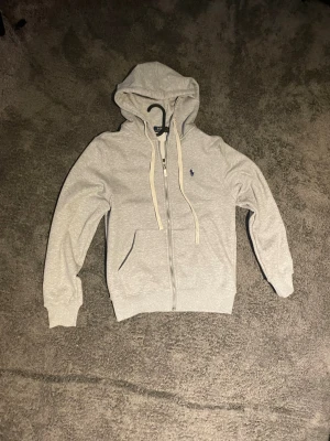 Ljusgrå hoodie med dragkedja - Snygg ljusgrå hoodie med dragkedja framtill och två fickor. Klassisk huva med vita snören och den ikoniska lilla blå loggan broderad på bröstet. Tillverkad i mjukt sweatshirtmaterial som är perfekt för chill dagar.