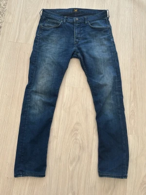  Lee Mörkblå slim fit jeans - Snygga mörkblå jeans i slim fit-modell med klassisk femficksdesign och lätt slitna detaljer på framsidan. Jeansen har normal midja och raka ben, perfekta för en avslappnad men stilren look. Materialet är troligtvis bomull med lite stretch för extra komfort.