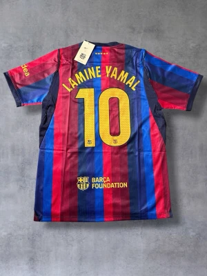 Barcelona matchtröja Lamine Yamal #10 - Snygg FC Barcelona matchtröja med Lamine Yamal och nummer 10 på ryggen. Tröjan har klassiska blå och röda vertikala ränder, gul Nike-logga och klubbmärke på bröstet samt Spotify-logga. Tillverkad i lätt och ventilerande material, perfekt för fotbollsträning eller match.