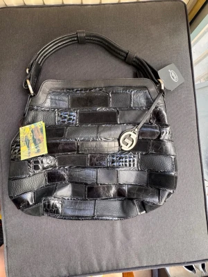 Svart patchwork handväska i skinn - Snygg svart handväska i patchwork-design med olika strukturer av skinn, inklusive blankt och krokopräglat läder. Väskan har ett brett handtag och en dekorativ metallberlock framtill. Perfekt för dig som gillar en unik och trendig look. Märket är gianni conti