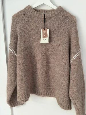 Beige stickad tröja från JDY, Helt oanvänd  - Mysig beige stickad tröja från JDY med rund halsringning och ribbade muddar. Tröjan har en avslappnad passform och vita dekorativa detaljer vid ärmarna. Perfekt för lager på lager och höstmys.