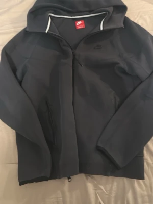 Svart hoodiejacka från Nike - Snygg svart hoodiejacka från Nike med dragkedja framtill och två sidofickor. Jackan har huva med vit kantdetalj och är tillverkad i ett mjukt material som känns sportigt. Perfekt för dig som gillar enkel och stilren design.