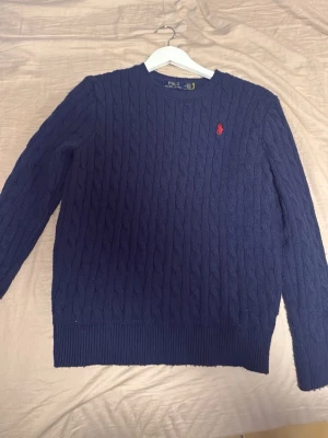 Mörkblå kabelstickad tröja Polo Ralph Lauren - Kabelstickad mörkblå tröja från Polo Ralph Lauren med rund halsringning och röd broderad logga på bröstet. Långa ärmar och ribbade muddar vid ärmslut och nederkant. Perfekt för lager på lager och klassisk stil.