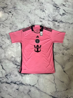 Rosa Inter Miami matchtröja Messi - Snygg rosa matchtröja från Inter Miami med svarta detaljer och Messi 10 på ryggen. Tröjan har korta ärmar, rund hals och klubbmärke samt Adidas-logga på bröstet. Tillverkad i lätt och ventilerande material, perfekt för fotboll eller träning.