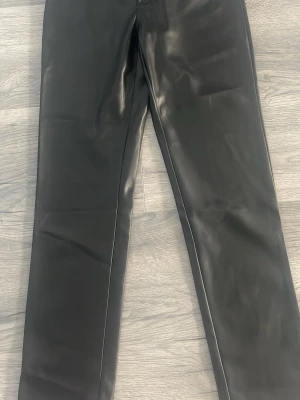 Svarta raka byxor i skinnimitation - Snygga svarta byxor i skinnimitation med rak passform och hög midja. Materialet har en glansig finish som ger en cool look. Perfekta för dig som vill ha ett trendigt och edgy plagg i garderoben. 36 midjan 32 storlek 