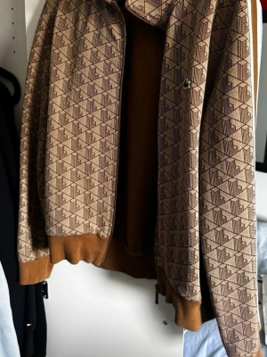 Beige mönstrad Lacoste ziptröja med revärer - Säljer en beige Lacoste ziptröja med brunt geometriskt mönster och vita/bruna revärer på ärmarna. Tröjan har ribbade muddar och hög krage, samt dragkedja framtill. Perfekt för en sportig och trendig look.