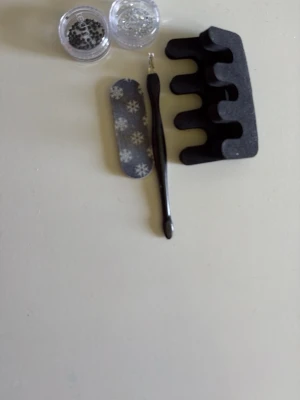 Nagelkit med dekorationer och verktyg - Ett komplett nagelkit med svart tåseparator i mjukt skum, svart nagelbandspetare, nagelfil med snöflingemönster samt två burkar med svarta och silvriga nageldekorationer. Perfekt för dig som vill fixa snygga naglar hemma.