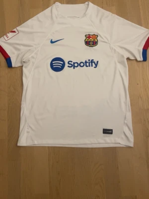 Vit FC Barcelona matchtröja Nike - Säljer en vit FC Barcelona matchtröja från Nike med klubbmärke och Spotify-logga på bröstet. Tröjan har blå och röda detaljer vid ärmsluten samt en liten Nike-logga i blått. Perfekt för dig som älskar fotboll och vill stötta Barca.
