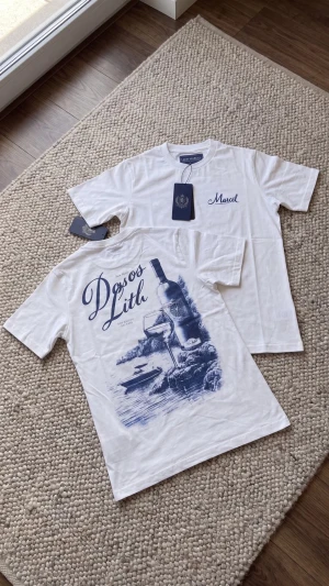Vit t-shirt med blått tryck - Vit t-shirt med blått tryck på ryggen som föreställer vinflaska, glas och landskap samt texten 'Dos os Lith'. Framtill finns en liten blå logga/text på bröstet. Klassisk passform och rund hals.