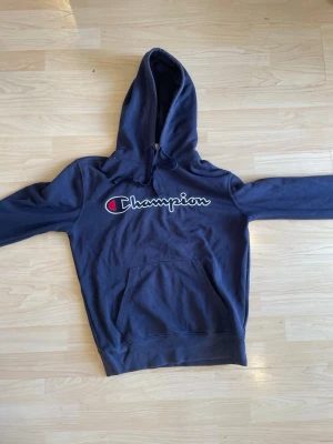 Mörkblå Champion hoodie med logga - Mörkblå hoodie från Champion med stor broderad logga över bröstet. Klassisk känguruficka framtill och justerbar huva med snören. Perfekt för en avslappnad och sportig stil. Material: bomull, polyester, elastan