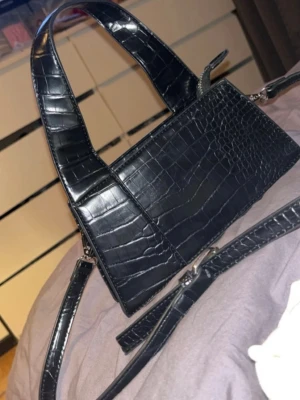 Svart croco-mönstrad handväska - Snygg svart handväska med croco-mönster och stilren rektangulär form. Väskan har både ett kort handtag och en avtagbar axelrem för olika stilar. Materialet är glansigt konstläder som ger en lyxig känsla. Perfekt accessoar för att lyfta din outfit.