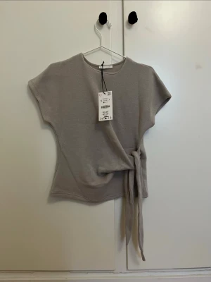 Snygg beige topp med knyt  - Snygg beige topp med korta ärmar och knyt i midjan som spänner till outfiten lite. Toppen har en enkel, stilren design och är tillverkad i ett mjukt material som känns skönt mot huden. Perfekt att styla med jeans eller kjol. Ny med prislapp!