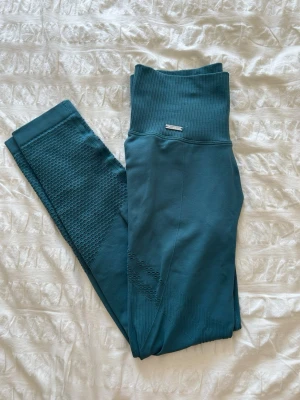 Turkosa seamless leggings med hög midja från amin - Säljer ett par turkosa seamless leggings med hög ribbad midja och diskret strukturmönster längs benen. Stretchigt material som sitter skönt och formar sig efter kroppen. Perfekta för träning eller chill. Liten metallbricka framtill vid midjan.