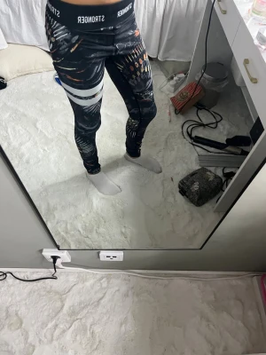 Svarta Stronger leggings med mönster - Säljer ett par svarta leggings från Stronger med färgglatt abstrakt mönster och vita ränder på ena benet. Med logotyp i midjebandet. Stretchigt material som sitter tight och bekvämt. Perfekta för träning eller chill.