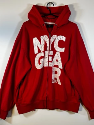 Oxide NYC Zip Hoodie Röd – XL / Y2K Streetwear - Röd zip hoodie från Oxide i storlek XL med stort vitt NYC-tryck framtill. Snygg oversized look med naturligt distressed/cracked print som ger en tydlig vintage/Y2K-känsla.  • Storlek XL, passar mer som L • Full zip • Stor huva • Stort NYC graphic print • Fickor framtill • Röd/vit färgkombination  Begagnat skick med normalt slitage, se bilder för skick.