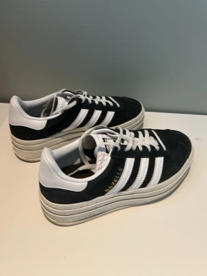Adidas Gazelle svartvita sneakers - Säljer ett par Adidas Gazelle sneakers i svart mocka med vita detaljer och klassiska tre ränder på sidan. Skorna har en chunky vit sula och vita skosnören. Perfekta för dig som gillar retrostil och vill sticka ut lite extra. Kom med frågor 😀