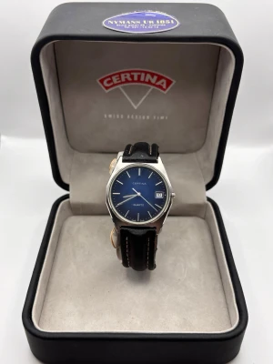 Certina klocka med svart läderband - Stilren Certina klocka med blå urtavla, datumvisning och silverfärgat boett. Klockan har ett klassiskt svart läderband med vita sömmar och en rund form. Perfekt accessoar för dig som gillar tidlös design och kvalitet.
