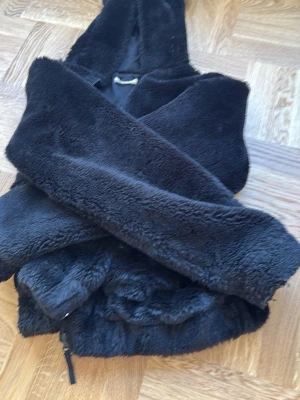 Svart teddyjacka med huva - Mysig svart teddyjacka med huva och dragkedja framtill. Jackan har ett fluffigt, mjukt material och en dekorativ träknapp som detalj. Perfekt för kyliga dagar när du vill vara både varm och stilren.
