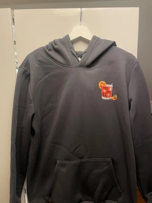 Grå hoodie med Negroni-tryck - Snygg grå hoodie med stor färgglad Negroni-illustration och text på ryggen samt mindre tryck på bröstet. Hoodien har huva och känguruficka framtill. Perfekt för dig som gillar coola drinkmotiv och vill sticka ut lite extra.
