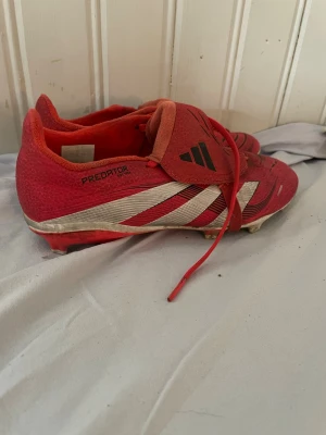 Adidas Predator röda fotbollsskor - Säljer ett par röda Adidas Predator fotbollsskor med vita och svarta detaljer. Skorna har klassisk snörning, låg profil och dobbar för spel på gräs. Tillverkade i syntetmaterial för bra grepp och kontroll på planen. Perfekta för dig som vill sticka ut på matchen.