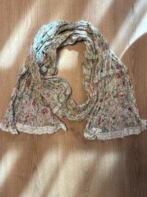 Blommig sjal med spetskant - Söt sjal i ljusbeige med småblommigt mönster i rosa, grönt och orange. Sjalen har en mjuk, plisserad struktur och avslutas med en vit spetskant som ger en romantisk touch. Perfekt accessoar för att lyfta din outfit.