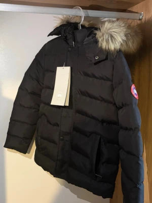 Svart Canada Goose jacka barn - Snygg svart jacka från Canada Goose med avtagbar päls. Jackan har klassisk patch på ärmen, dragkedja och knappar framtill samt två stora fickor. Perfekt för kalla vinterdagar och riktigt varm. Flera storlekar finns!!!