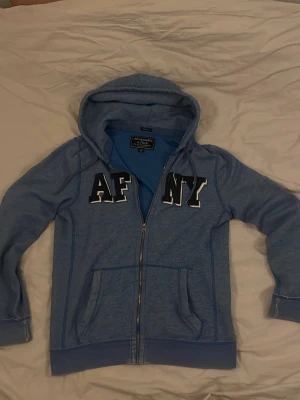 Abercrombie & Fitch hoodie  - Riktigt fet blå zip-hoodie från Abercrombie & Fitch med stora vita och svarta bokstäver 'AF NY' på bröstet. Hoodien har dragkedja, två fickor fram och en klassisk huva. Perfekt för en avslappnad och cool stil. Storlek M. 