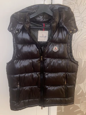 Svart dunväst från Moncler - Snygg svart dunväst från Moncler med glansig finish, dragkedja framtill och två fickor med dragkedja. Västen har huva och Moncler-logga på bröstet. Perfekt för lager-på-lager och streetstyle.