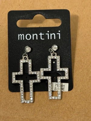 Korsörhängen med strass från Montini - Säljer ett par silvriga örhängen i form av kors från Montini. Örhängena är dekorerade med gnistrande strass-stenar längs hela kanten och har en klassisk krok. Perfekta för dig som vill sticka ut med en cool accessoar.