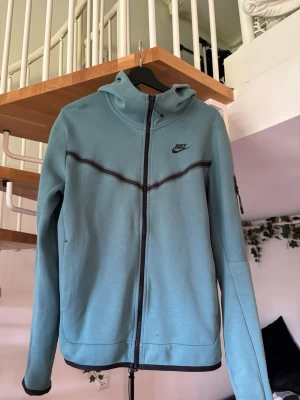 Blå zip hoodie från Nike - Snygg ljusblå hoodie från Nike med dragkedja framtill och svart Nike-logga på bröstet. Hoodien har huva, svarta detaljer längs dragkedjan och ett diskret svart band över bröstet. Tillverkad i mjukt material, perfekt för chill dagar.