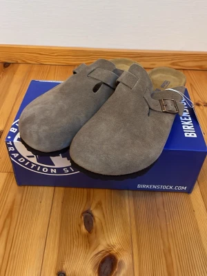 Grå mockatofflor från Birkenstock - Säljer ett par gråa tofflor från Birkenstock i mjuk mocka. Modellen har täckt tå, justerbart spänne i metall och klassisk korksula för extra komfort. Perfekta för dig som gillar stilrena och bekväma skor till vardags.