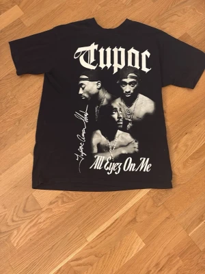 Svart Tupac t-shirt Pull&Bear - Svart t-shirt från Pull&Bear med stort Tupac-tryck och texten 'All Eyez On Me' framtill. Oversized passform och klassisk rund hals. Perfekt för dig som gillar streetwear och hiphop-vibbar.