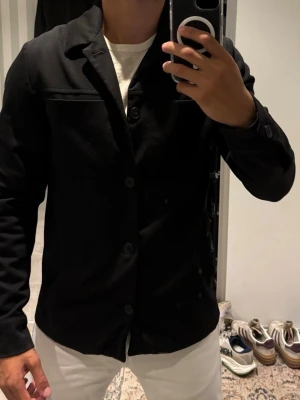 Svart overshirt från Les Deux - Stilren svart overshirt från Les Deux med klassisk krage och knäppning framtill. Två bröstfickor och knappdetaljer vid ärmsluten ger en clean look. Perfekt att bära över en t-shirt för en avslappnad men snygg stil.