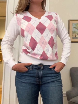 Vit och rosa argyle stickad tröja - Snygg stickad tröja med klassiskt argylemönster i rosa, vitt och ljusblått. Tröjan har v-ringning, långa ärmar och ribbade muddar. Perfekt för en preppy look och passar till jeans eller kjol.
