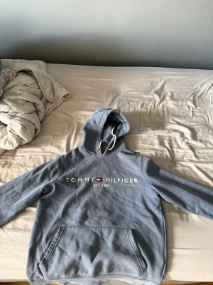 Blå hoodie från Tommy Hilfiger storlek S - Snygg blå hoodie från Tommy Hilfiger med broderad logga och text 'EST. 1985' på bröstet. Klassisk känguruficka framtill och justerbar huva med vita snören. Perfekt för en avslappnad och trendig stil storlek S