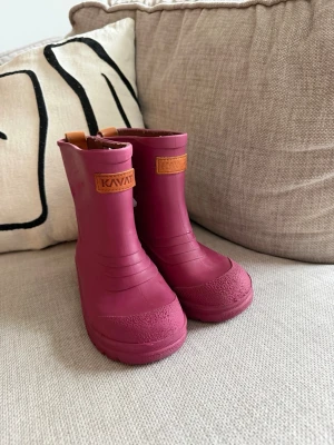 Rosa gummistövlar från Kavat - Säljer ett par rosa gummistövlar från Kavat med brunt logomärke framtill. Stövlarna är tillverkade i slitstarkt gummi och har en klassisk, rundad tå och förstärkt tåparti. Perfekta för regniga dagar och lek utomhus.