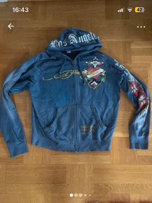 Blå Ed Hardy hoodie med tryck och nitar - Säljer en blå zip hoodie från Ed Hardy med stora tryck både fram och bak, samt coola detaljer som nitar på fickorna och blommor på ena ärmen. Huvan har texttryck och materialet känns mjukt och bekvämt. Perfekt för dig som gillar streetwear och unika plagg.