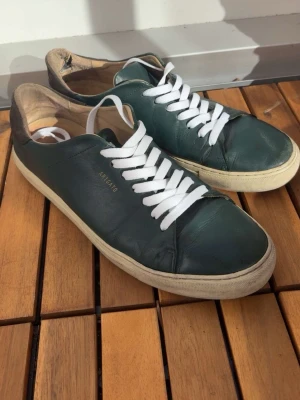 Mörkgröna sneakers från Axel Arigato - Snygga mörkgröna sneakers från Axel Arigato med vita snören och beige sula. Skorna är i läder och har en stilren, minimalistisk design med diskret logga på sidan. Perfekta för dig som gillar klassiska och cleana sneakers.