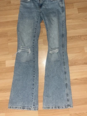 Ljusblå bootcut jeans med slitningar - Säljer ett par ljusblå bootcut jeans med slitningar över knäna och råa fållar. Jeansen har klassisk femficksdesign och är tillverkade i denim. Perfekta för en avslappnad och trendig look.